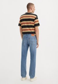 Gestreepte shirt met korte mouwen in zwart, rood en geel, gecombineerd met lichtblauwe rechte spijkerbroek en crème sneakers, gezien vanaf de achterkant.