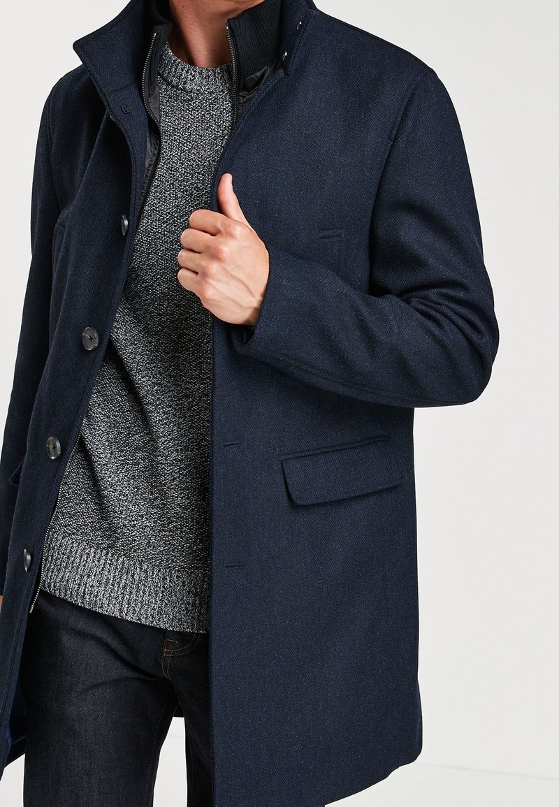 Homme portant un manteau bleu foncé boutonné sur un pull en maille gris et un jean foncé, tenant le col du manteau d'une main.