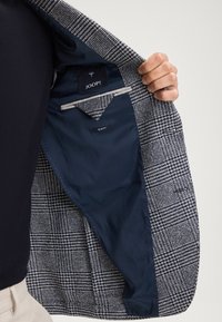 Blazer con motivo a quadri blu e bianchi, foderato in tessuto blu. L'etichetta riporta "JOOP!" e "SLIM FIT." Taschino interni visibili.