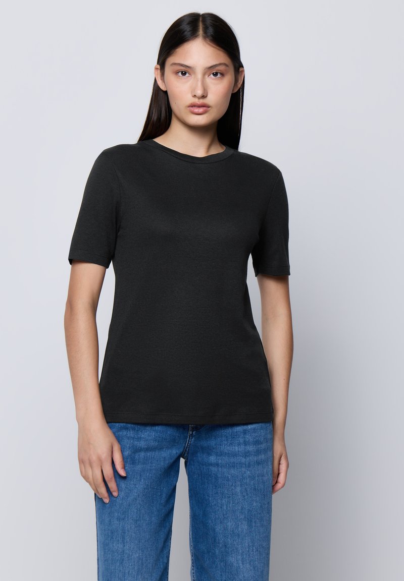 Giovane donna con lunghi capelli scuri che indossa una semplice maglietta nera a maniche corte e jeans blu, in piedi di fronte a uno sfondo grigio chiaro.