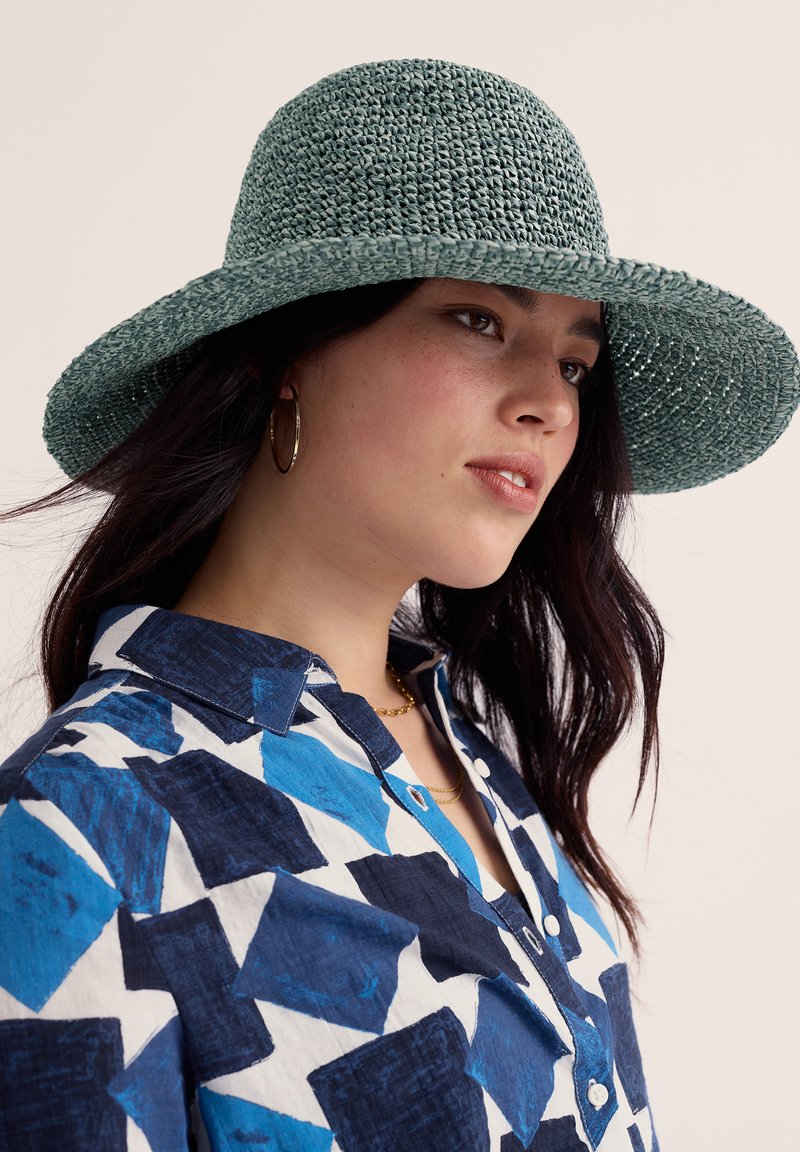 Noor naine tumedate juustega, kellel on peas roheline punutud lai äärega kübar, kuldsed rõngakujulised kõrvarõngad ning sinine geomeetrilise mustriga kraega särk.