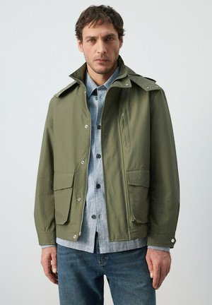 Hombre con una chaqueta verde con cremallera sobre una camisa gris abotonada y vaqueros azules, de pie contra un fondo claro y sencillo.