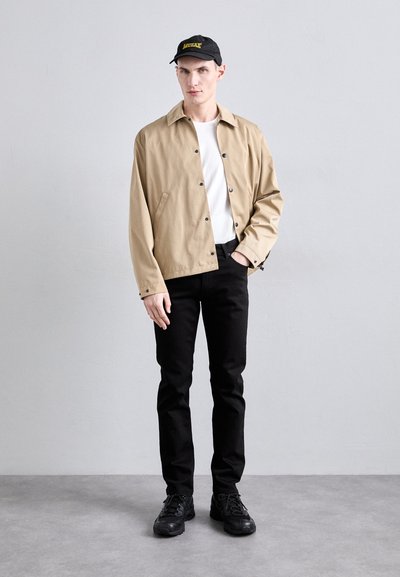 Beige leichte Jacke mit Knöpfen, weißes T-Shirt, schwarze Hose und schwarze Sneaker. Schwarze Mütze mit gelbem Logo-Detail.