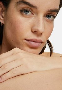 Närbild av ett ansikte med klar hy, blå ögon och subtil sminkning. Guldörhängen med en blå sten. Ljus hy och ljusbrunt hår.