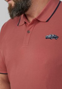 Bärtiger Mann, der ein rotes Poloshirt mit marineblauen Akzenten und einem kleinen bestickten navyblauen Vintage-Auto auf der Brust trägt.