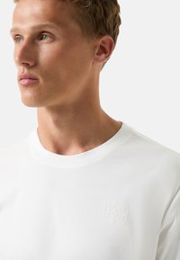 Hvid bomulds T-shirt med rund halsudskæring og et lille, præget logo på brystet. Glat tekstur og afslappet pasform.
