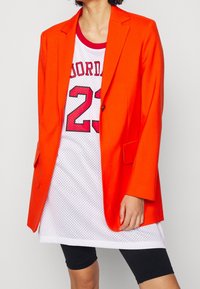 Orange skräddarsydd blazer med kavajslag över en vit meshtröja med röda detaljer och svarta tighta shorts. Slät textur.