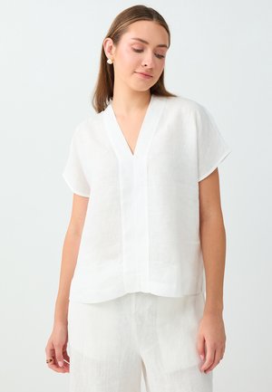 Jeune femme portant un haut blanc ample à manches courtes avec col en V et un pantalon blanc assorti, debout devant un fond clair uni.