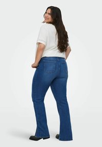 Jeans a zampa d'elefante blu scuro con una texture liscia, dotati di tasche posteriori. Il modello indossa una maglietta bianca a maniche corte con dettagli ad occhio di pernice.