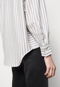 Chemise à manches longues à rayures verticales grises et blanches, avec un ourlet légèrement arrondi et des poignets boutonnés, associée à un pantalon noir.