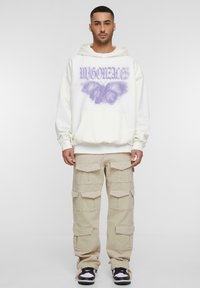 Hvid hættebluse med lilla sommerfugl grafisk design og tekst, parret med beige cargo bukser med flere lommer og sorte sneakers.