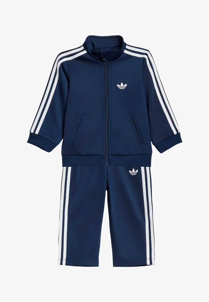 Ensemble de survêtement bleu marine pour tout-petit avec des bandes blanches Adidas sur les manches et les jambes, veste zippée et poignets élastiques.