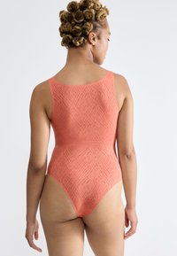 Vrouw met krullend haar in een mouwloze, koraalkleurige, gestructureerde body die met haar rug naar de camera staat, tegen een effen lichte achtergrond.