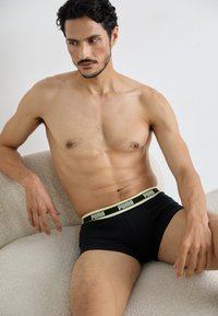 Fekete boxeralsó fehér és neon sárga Puma logóval a derékrészen, testhezálló fazon, amelyet egy férfi modell visel, miközben egy textúrált felületen ül.