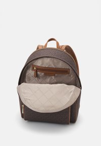 Brun quiltet stof rygsæk med læderdetaljer, der har en toplynlås, en forlommer og et rummeligt beige indre med diamantmønster.