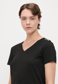 Sort V-hals t-shirt med en spættet tekstur, der har tre dekorative guldfarvede forskydninger på venstre skulder. Korte ærmer.