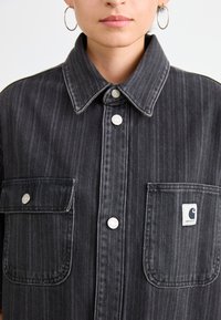 Schwarzes Denim-Hemd mit vertikalen Streifen, großem Kragen, silbernen Beschlägen, zwei Brusttaschen und einem kleinen Logo-Patch auf der rechten Tasche.