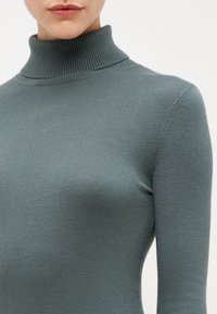 Ribbet genser med høy hals i dempet teal. Laget av myk, elastisk stoff, kroppsnær design, lange ermer, glatt tekstur.