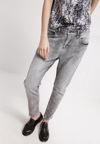 Jeans de mezclilla grises con corte ajustado, que cuentan con bolsillos frontales y un cierre de botón, combinados con zapatos negros de cordones.