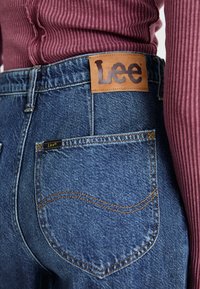 Blue denim jeans with a leather Lee brand tag. Features back pocket stitching and a snug fit. Accompanied by a ribbed maroon top.