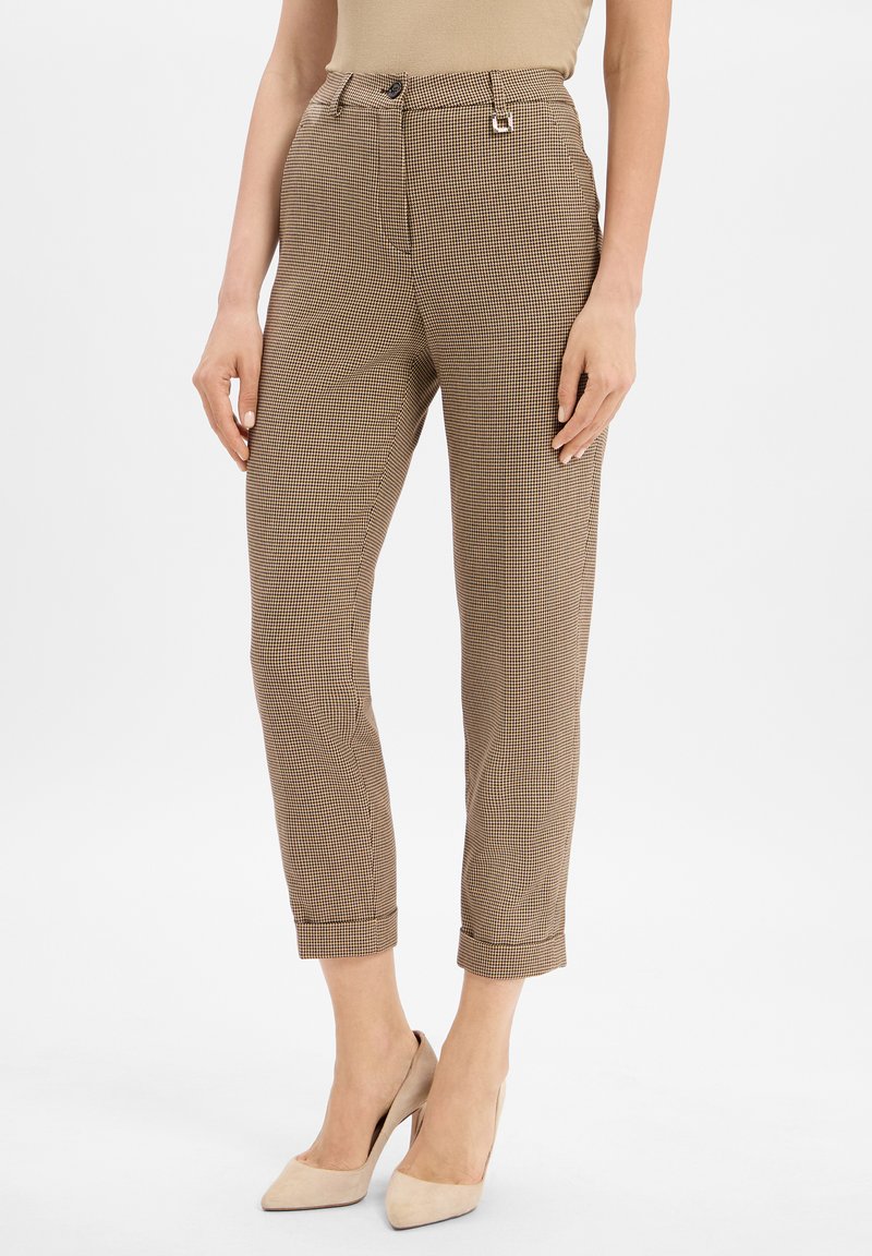 Femme portant un pantalon à carreaux beige et marron coupe cheville avec des talons hauts beiges, se tenant devant un fond blanc.