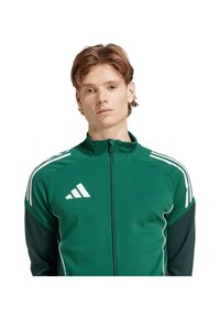 Giacca atletica verde con zip, strisce bianche e logo. Realizzata in tessuto liscio, design aderente, con maniche in contrasto.