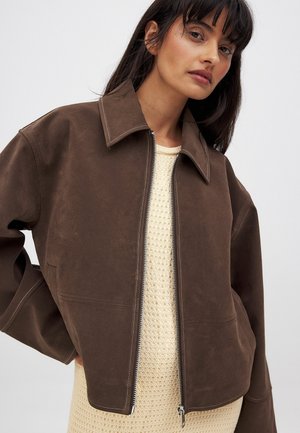 Brun ruskindjakke med notched krave, lynlås lukning og hvide sydetaljer, lagdelt over en tekstureret beige top.