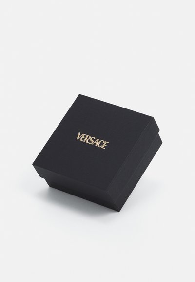 Boîte en carton noire avec une surface texturée, portant le logo doré "VERSACE" sur le dessus. Forme carrée avec un design à couvercle pivotant.