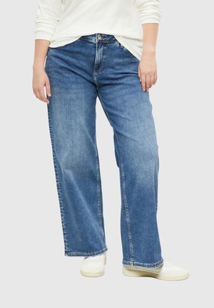 Person trägt hellblaue Jeans mit weitem Bein, weiße Sneakers und ein langärmliges weißes Oberteil vor einfarbigem Hintergrund.