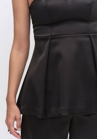 Musta peplum-toppi sileässä kankaassa, jossa on olkaimeton muotoilu. Luonnollinen vartalonmyötäinen yläosa ja levenevä helma. Ei näkyviä kiinnityksiä tai koristeluja.