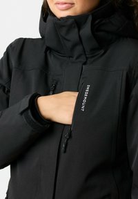 Black fleece-lined jacket featuring a high collar, zippered pockets, Velcro cuffs, and branded detail on the front. Smooth matte finish.