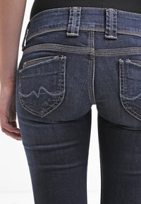 Tmavě modré džíny z denimu s úzkým střihem, které mají dvě zadní kapsy a kontrastní švy, zdůrazňující jednoduchý vlnkový vzor na kapsách.