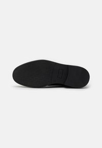 Suela de zapato negra con patrón texturizado, logo rectangular en el centro y detalle de tacón redondo. Material de goma con una superficie lisa.