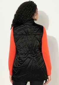 Černá prošívaná vesta s vysokým límcem, s geometrickým prošíváním. Nositá přes jasně oranžové dlouhé tričko. Pohled ze zadu.