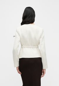 Blazer blanc texturé à manches longues avec une taille cintrée, agrémenté d'un détail ceinture. Associé à une jupe marron côtelée ajustée.