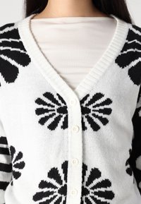 Cardigan noir et blanc avec des motifs floraux, décolleté en v, et cinq boutons, fabriqué en tissu tricoté doux avec des bordures côtelées.