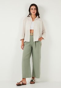 Kevyt, teksturoitu beige paita valkoisen crop-topin päällä, yhdistettynä löysiin, vaaleanvihreisiin housuihin ja koristeellisiin sandaaleihin, joissa on kukkakoristeita.