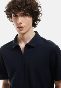 Mørkeblå shortsleeve polo t-shirt med lynlåskrage, udstyret med et tekstureret stof med vaffelmønster. Nærbillede af tøjet.