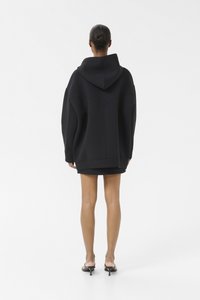 Zwarte oversized hoodie met een grote capuchon, ribgebreide manchetten en zoom, gemaakt van een gestructureerde stof. Draagt over een kortere zwarte jurk.