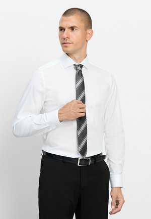 Mann richtet gestreifte schwarze und graue Krawatte, trägt weißes Hemd und schwarze Hose vor einem einfarbigen weißen Hintergrund.