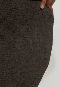 Strukturierter, dunkelbrauner Stoff mit einer zerknitterten Oberfläche. Die Nahaufnahme zeigt ein tailliertes Design und einen leichten Glanz im Material. Keine sichtbare Hardware.