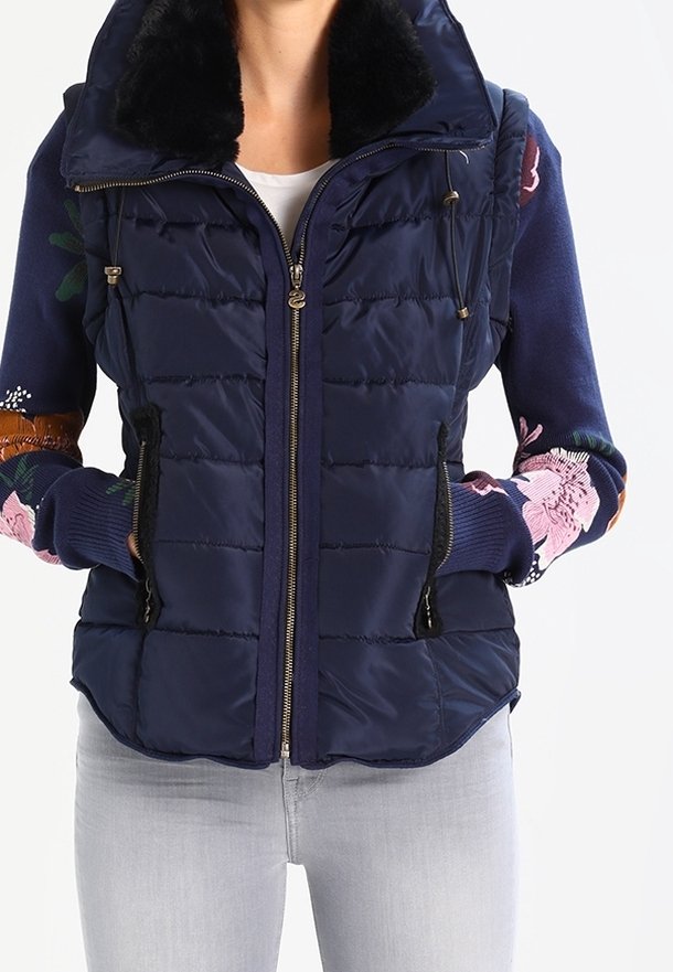 Femme portant une veste matelassée bleu marine avec une fermeture éclair et un col en fourrure noire par-dessus un pull à fleurs à manches longues et un pantalon gris clair.