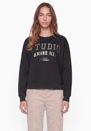 Ung kvinde med bølget brunt hår bærer en sort sweatshirt med hvid tekst og lyse beige bukser, står mod en ensfarvet hvid baggrund.