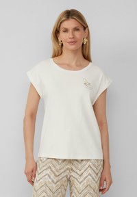 T-shirt en coton blanc avec des manches retroussées, arborant une petite broderie dorée en forme d'abeille sur le côté gauche. Le tissu a une texture douce et lisse.