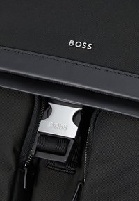 Juodas nailono kuprinės dizainas yra elegantiškas, sidabriniai akcentai su "BOSS" įspaudu ir tekstūruotas paviršius. Matomi užtrauktukai.