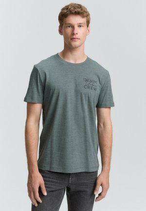 Fiatal férfi rövid szőke hajjal, zöld, kerek nyakú pólót visel, amelyen a "Denim Urban Crew" felirat áll, sötét farmernadrággal párosítva, egy egyszínű háttér előtt áll.