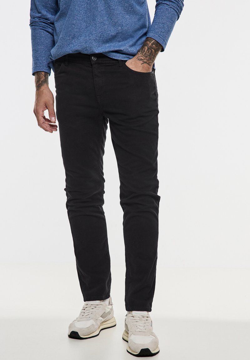 Schwarze Baumwoll-Chinohosen im Slim-Fit, mit Vorderfächern und einer subtilen Textur, kombiniert mit hellen Sneakern.