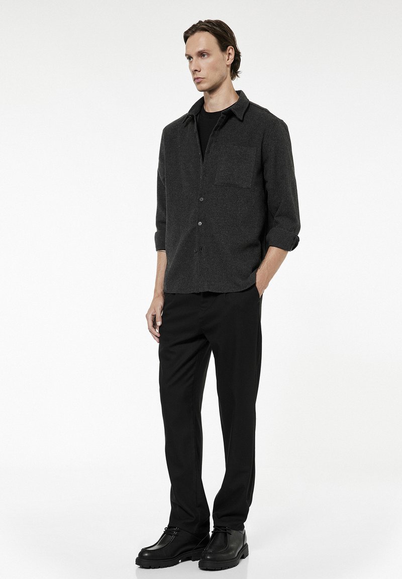 Homme portant une chemise boutonnée gris foncé avec les manches retroussées, un pantalon noir et des chaussures noires, debout devant un fond blanc uni.