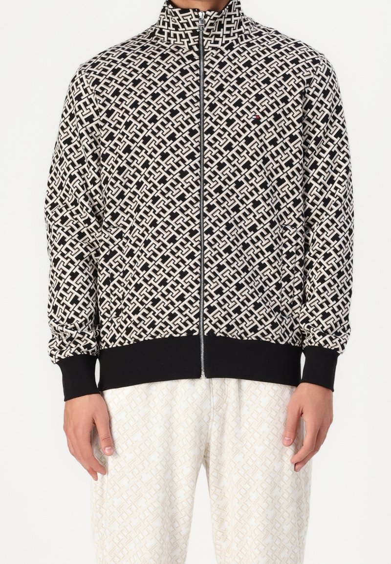 Homme portant une veste zippée noire et blanche à motifs avec des poignets noirs et un pantalon blanc à motifs avec un design géométrique subtil.