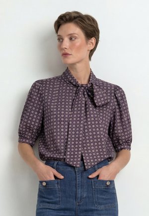 Mujer con cabello corto que lleva una blusa morada con estampado, cuello con lazo y vaqueros azul de talle alto con las manos en los bolsillos.